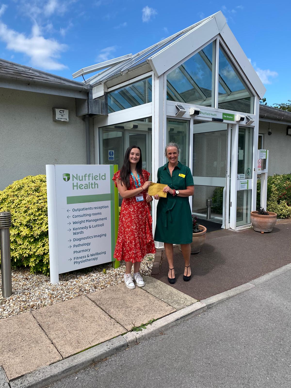 Two women standing outside Nuffield Health holding an envelope as part of St Margaret’s Hospice’s Charity £50 Challenge for businesses fundraising campaign.